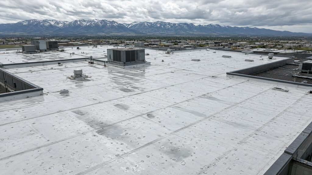 Wet white commercial membrane roof with hail impact marks and rooftop units, Front Range mountains in the background